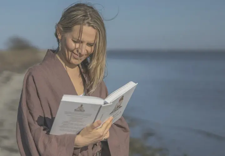 Kobieta w brązowym kimonie czyta książkę na plaży, z morzem w tle.