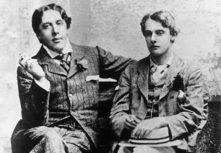 Oscar Wilde, lord elegancji i błyskotliwy pisarz, siedzi z młodym mężczyzną, trzymając papierosa.