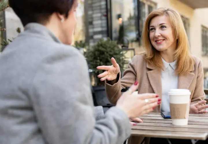 Dwie kobiety rozmawiają przy stoliku, gestykulując. Jedna z nich, z blond włosami, uśmiecha się, opowiadając o swoich przemyśleniach na temat granic w związku.