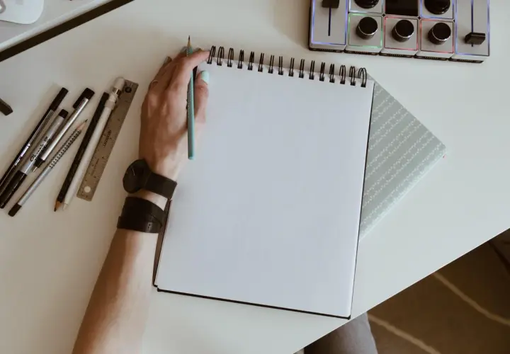 Mano con lápiz azul lista para crear dibujos de cumpleaños en un cuaderno blanco. A su lado, reglas, rotuladores y un reloj.