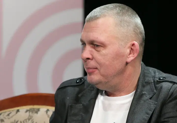 Mężczyzna z krótkimi włosami, w szarej kurtce, siedzi na kanapie. To wokalista zespołu Raz Dwa Trzy.
