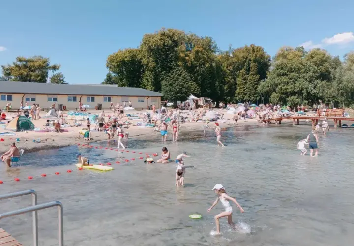 Słoneczny dzień na powidz plaży. Ludzie opalają się, kąpią i bawią w wodzie. W tle budynek i drzewa.