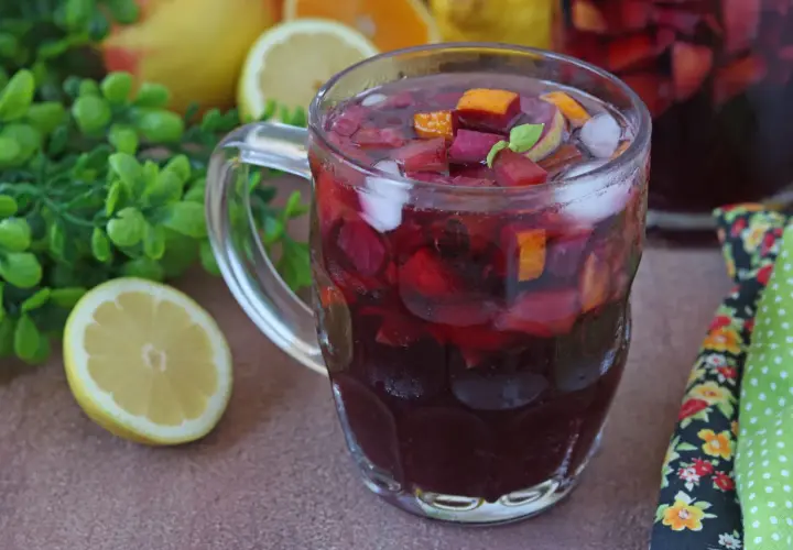 Un bicchiere colmo di sangria, con pezzi di frutta rossa, arancia e ghiaccio. La ricetta perfetta per l'estate, con limoni e foglie verdi sullo sfondo.