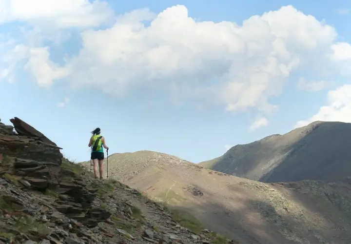 Senderista en la ruta Vall de Núria, con mochila y bastones, ascendiendo por un sendero rocoso con montañas al fondo.