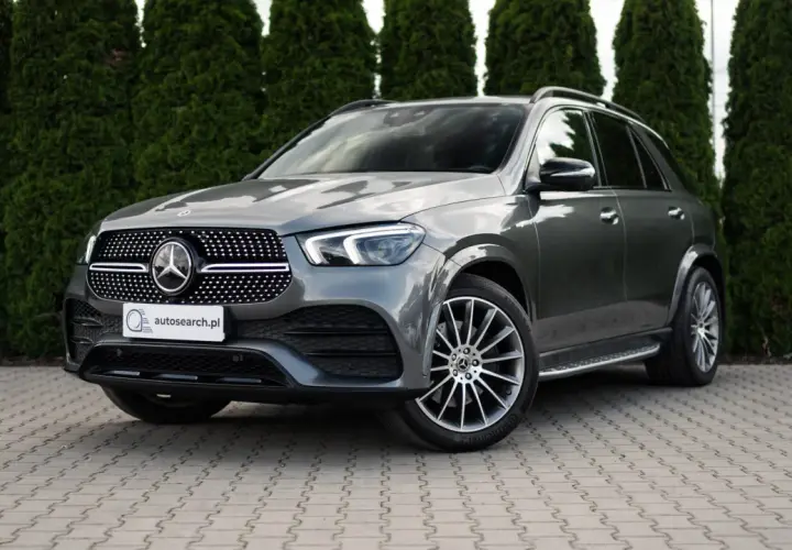 Elegancki, szary Mercedes GLE 400d SUV stoi na wybrukowanym podjeździe, otoczony gęstymi, zielonymi żywopłotami.