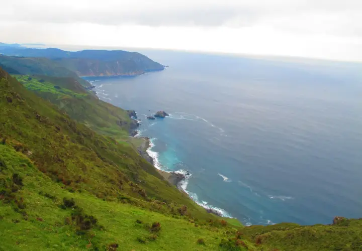 Paisaje costero verde y escarpado cerca de la garita de Herbeira, con el mar azul y las olas rompiendo en la costa rocosa.
