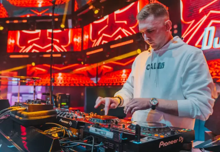 DJ w białej bluzie z kapturem gra na konsoli DJ-skiej na tle czerwonych świateł scenicznych.