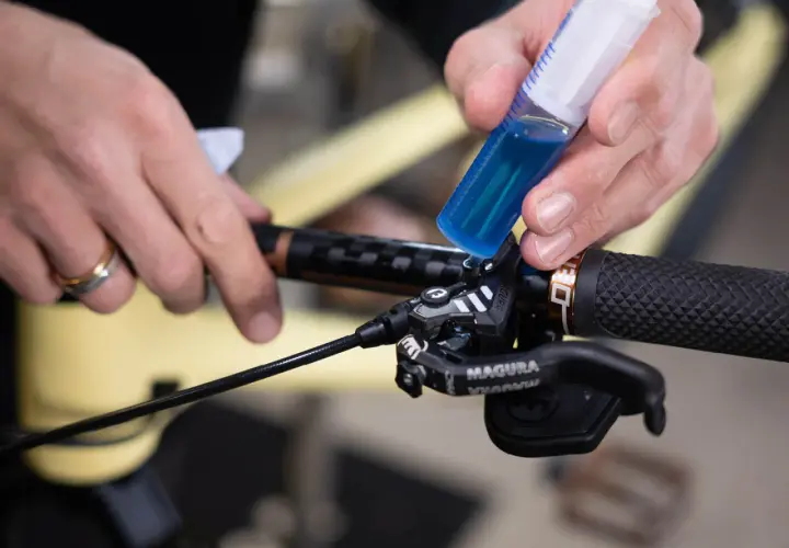 Hands add blue brake fluid from a syringe to a bicycle brake lever. This fluid is corrosive due to its hygroscopic nature, absorbing moisture.