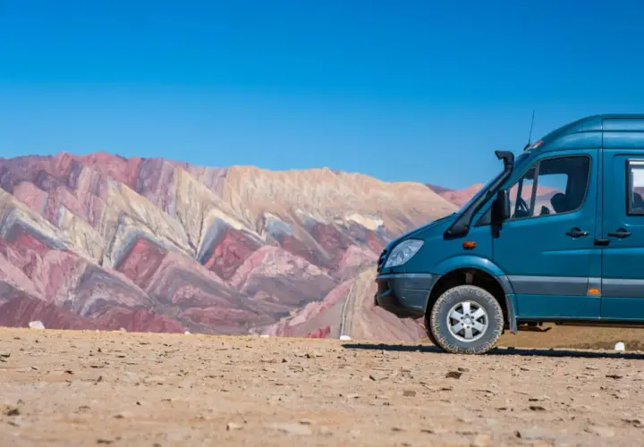 Un camping car tout terrain bleu est garé sur un sol rocailleux, avec en arrière-plan des montagnes aux couleurs vives et un ciel bleu éclatant.