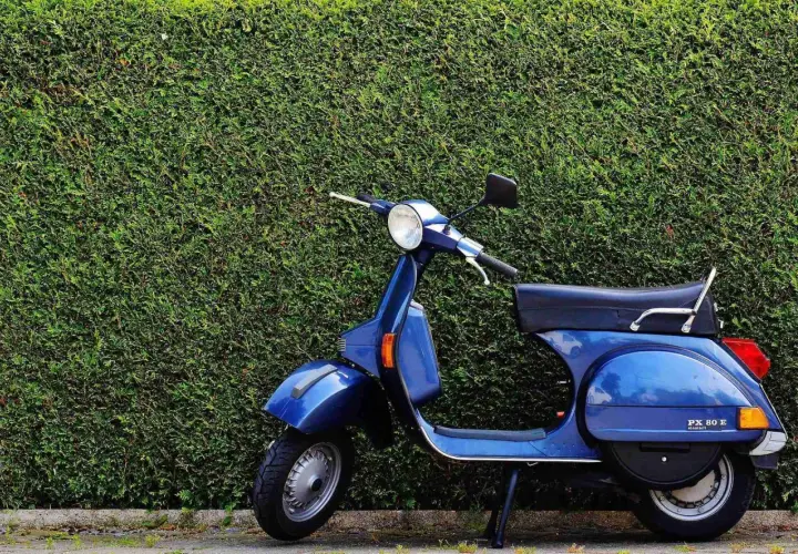 Azul Vespa PX 80 E, un clásico que evoca los modelos Vespa años 60, aparcada frente a un seto verde.
