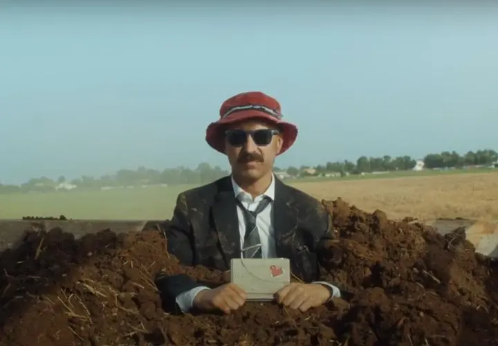 Uomo in abito e cappello rosso, sepolto nel terreno, tiene una lettera con un cuore. Una scena surreale per la recensione "il migliore dei mondi".