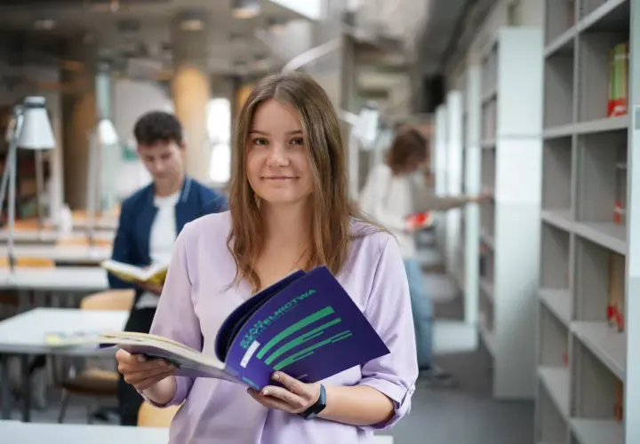 Studentka z uśmiechem trzyma broszurę, być może dotyczącą stypendium socjalnego. W tle inni studenci w bibliotece.