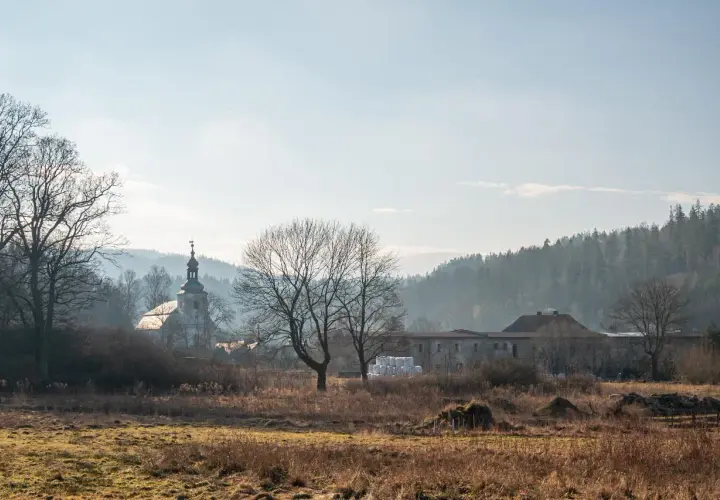 Krajobraz z kościołem i zabudowaniami, w tle las. W oddali widać mgliste wzgórza, charakterystyczne dla rudaw janowickich.