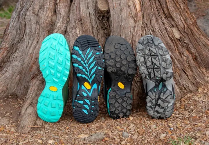 Four hiking boot soles lean against a tree. From left to right: bright teal, black with teal accents, black with circular lugs, and grey with a rugged tread. These sneaker soles are hard and slippery, designed for grip.