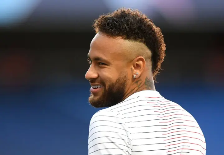 Sonrisa de Neymar con su característico peinado mohicano, luciendo una camiseta a rayas blancas y rojas.
