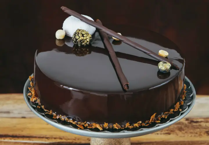 Torta cioccolatosa goduriosa con glassa lucida, decorata con riccioli di cioccolato, nocciole e una sfera dorata.