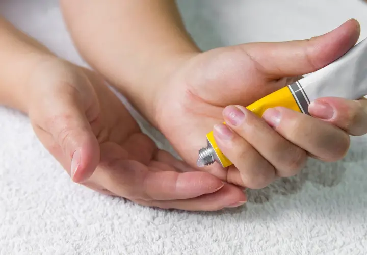 Manos aplicando crema de un tubo amarillo en la palma de otra mano.