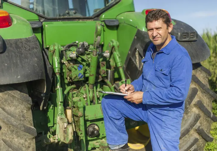 Bauer in blauer Latzhose prüft Unterlagen neben seinem Traktor. Er plant die nächste Ernte und denkt über den **Einkommensteuer Landwirtschaft** nach.