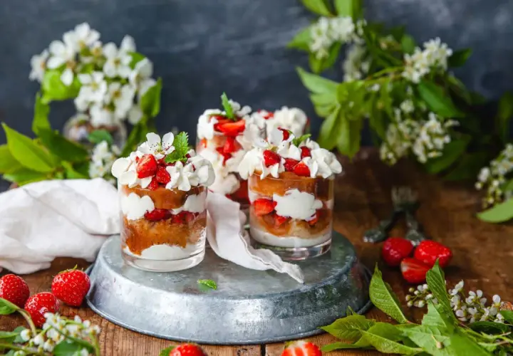 Tre glas med tårta jordgubbar, grädde och färska jordgubbar. Vårligt dekorerat med blommor och gröna blad.