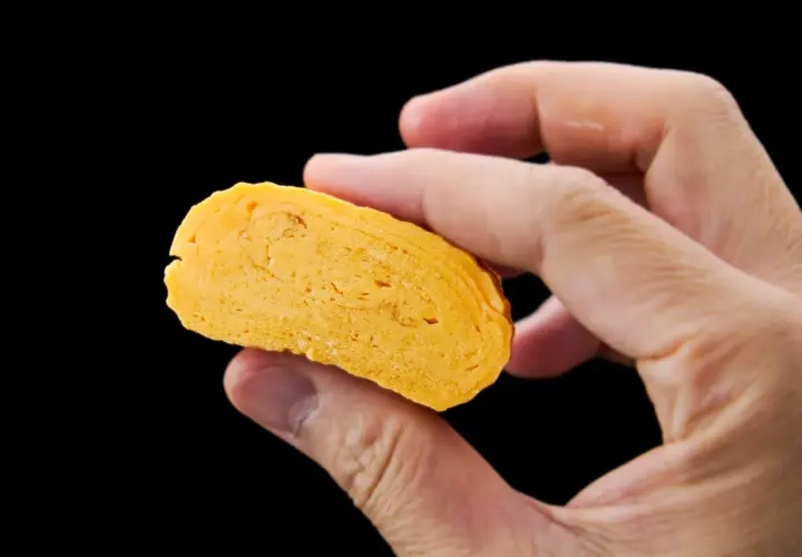 A hand holds a slice of golden-yellow dashimaki tamago, revealing its layered, fluffy texture.