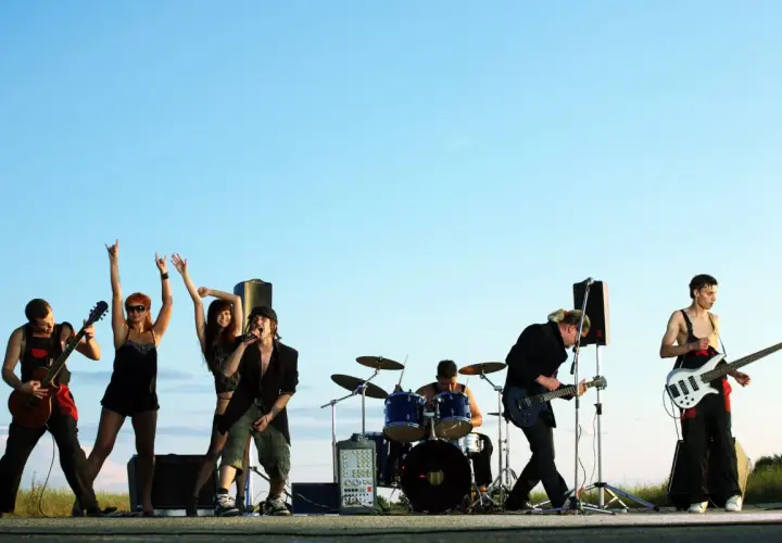 Zespół rockowy na tle nieba. Wokalista, gitarzysta, basista, perkusista i dwie tancerki. Zespoły rockowe zagraniczne alfabetycznie.