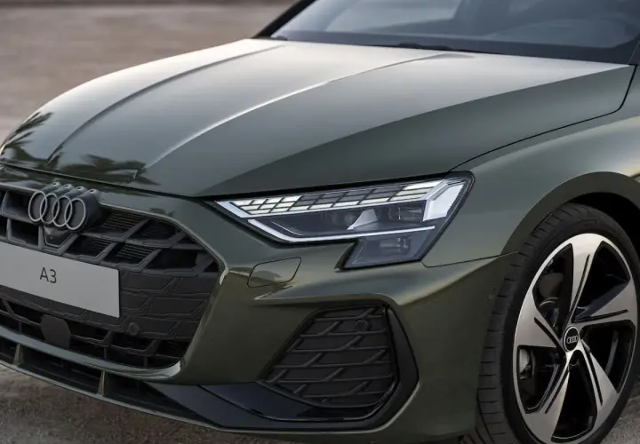 Nowe Audi A3 w zielonym kolorze prezentuje się elegancko. Widoczny fragment maski, grill z logo i reflektor podkreślają jego nowoczesny design.
