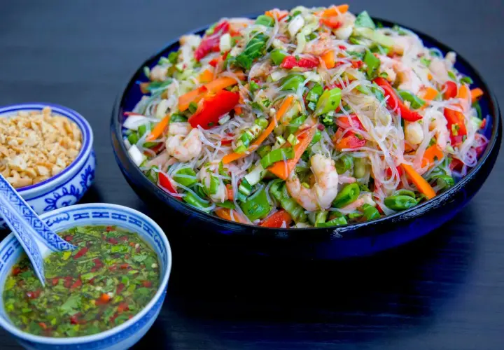 En stor skål med färgglad glasnudel sallad med räkor, paprika och grönsaker. Serveras med en dressing och hackade nötter.