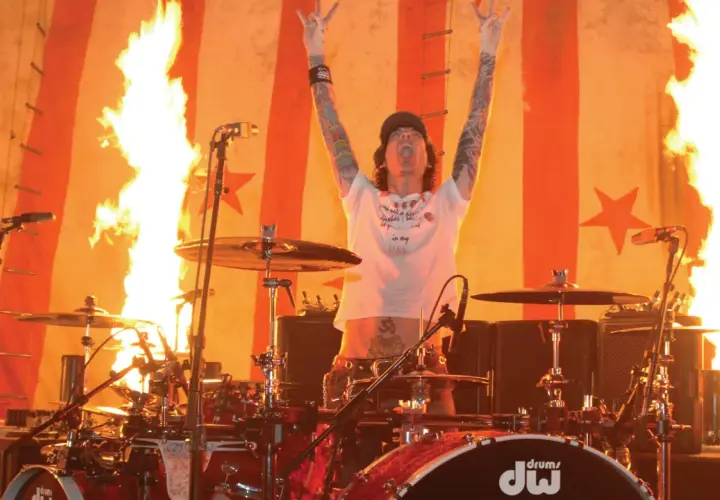 Tommy Lee w ogniu, z rękami w górze i pałeczkami perkusyjnymi, na tle płonącej sceny.