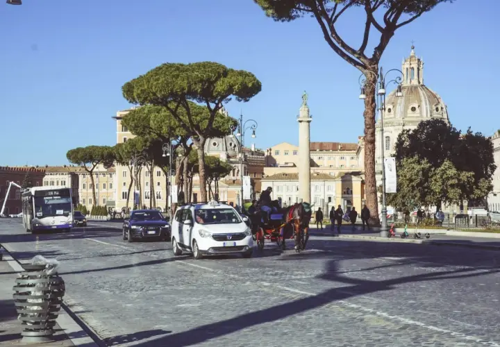 Carrozza trainata da cavalli, taxi e autobus sfilano su un lastricato romano, con la cupola di una chiesa e un monumento antico sullo sfondo, nella zona Battistini Roma.
