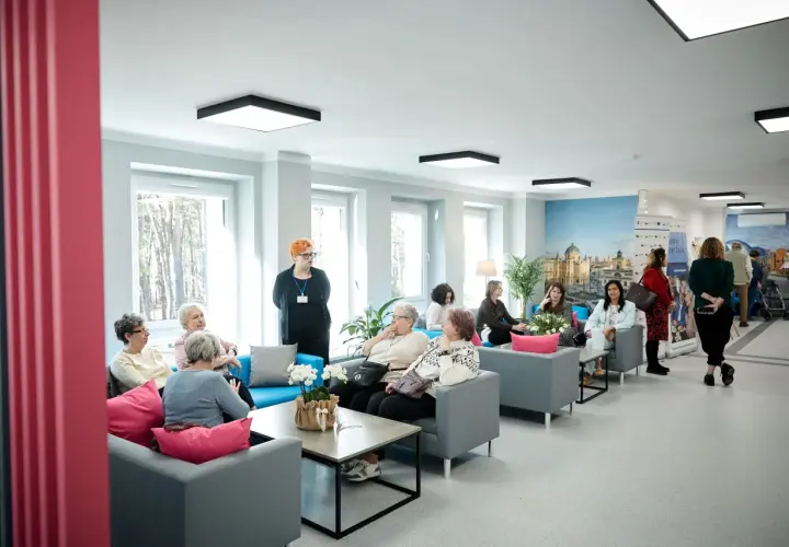 Grupa seniorów siedzi na kanapach w jasnym, nowoczesnym wnętrzu domu opieki.