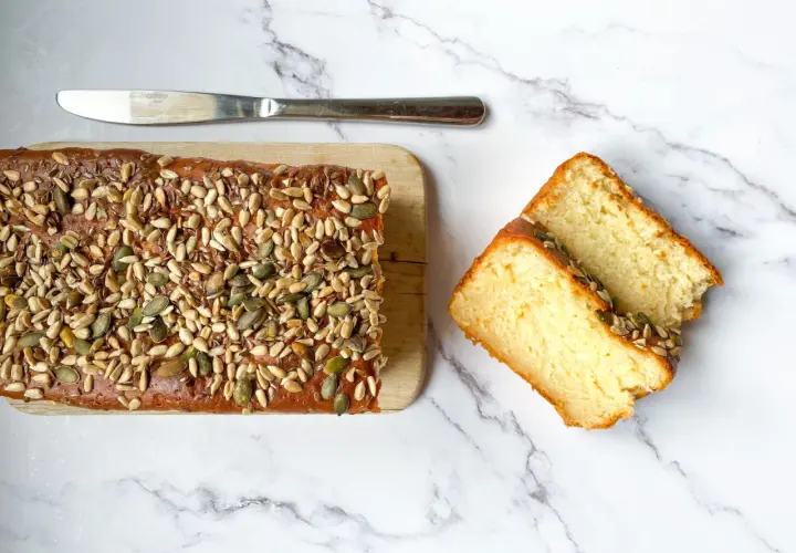 Plumcake senza uova, soffice e dorato, ricoperto di semi misti. Due fette tagliate mostrano la sua morbidezza.