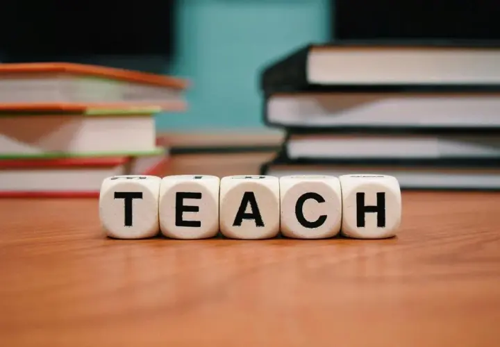 Klocki z literami "TEACH" symbolizują naukę i kolejność wprowadzania liter w klasie 1. W tle stosy książek.
