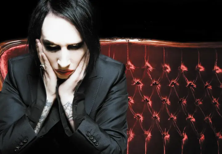 Marilyn Manson, z charakterystycznym makijażem, siedzi na aksamitnej kanapie. Jego utwory często poruszają mroczne tematy.