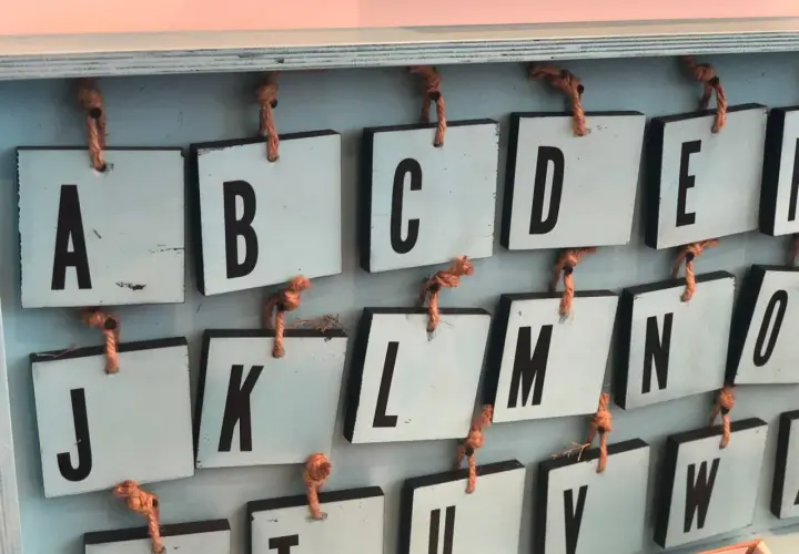 Drewniane tabliczki z literami alfabetu angielskiego pisanego, wiszące na sznurkach. Widoczne litery A, B, C, D, E, J, K, L, M, N, O, U, V, W.
