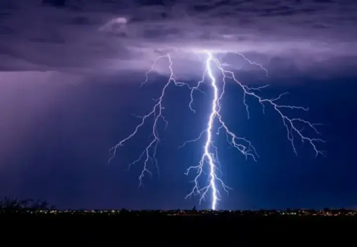 Un rayo ilumina el cielo nocturno, como si fuera un sueño con tormenta. Las nubes púrpuras y el horizonte iluminado por luces de ciudad crean una escena dramática.
