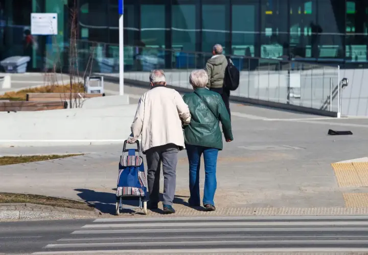 Starsza para idzie przez pasy, mężczyzna ciągnie wózek. To obraz codzienności, gdzie każdy krok to dowód na to, że życie trwa, a stopa zastąpienia nie jest jeszcze potrzebna.