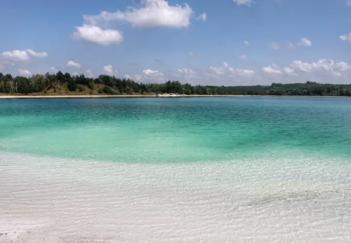 Lazurowe jezioro z piaszczystą plażą i lasem w tle. Woda jest krystalicznie czysta, a dno widoczne.
