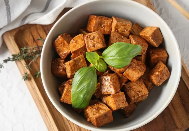 A bowl of glazed, cubed japanese tofu, garnished with fresh basil leaves, sits on a wooden tray next to chopsticks.
