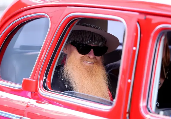 Billy Gibbons, jeden z członków ZZ Top, w stylowym kapeluszu i z charakterystyczną brodą, siedzi w czerwonym samochodzie.