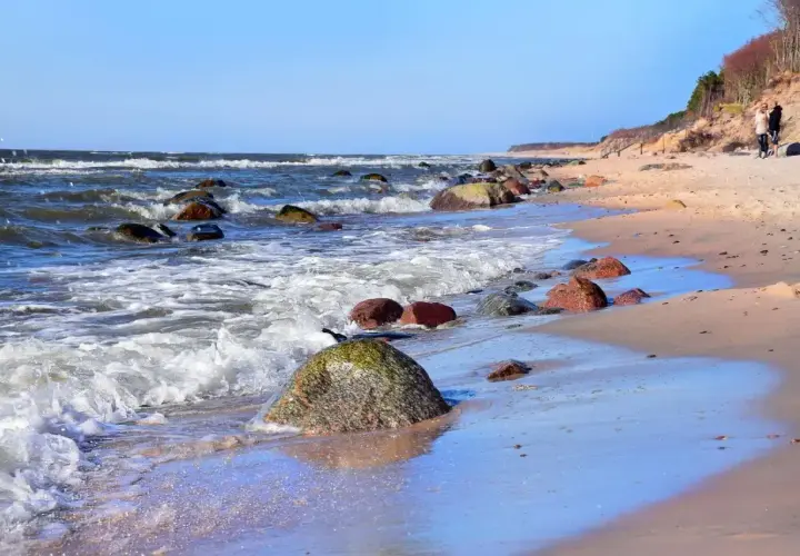 Poddąbie plaża. Fale rozbijają się o kamienie na piaszczystym brzegu. W oddali widać spacerującą parę.