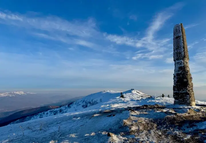 Kamienny obelisk na szczycie ośnieżonej góry, przypominający wielką rawkę. W tle błękitne niebo z chmurami i pasma górskie.