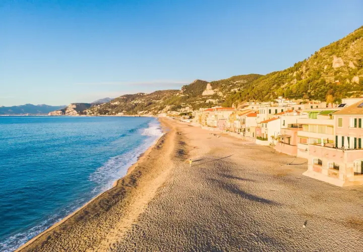 Plage de sable fin bordée de maisons colorées sur la côte italienne. La mer bleue et les collines verdoyantes complètent ce paysage paisible.