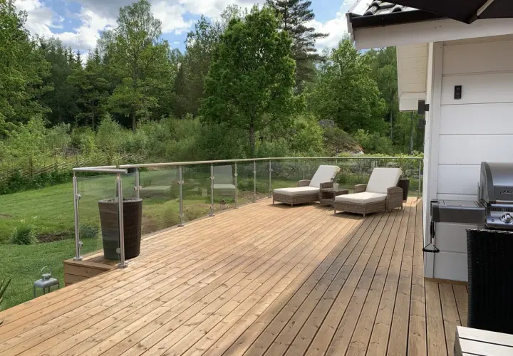 Träaltan med solstolar och grill, omgiven av grönskande natur. Ett nymålat staket syns i bakgrunden.
