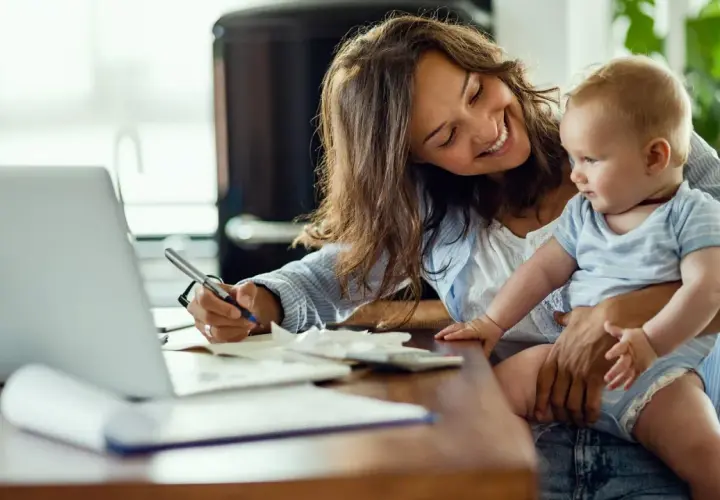 Lächelnde Mutter mit Baby auf dem Arm schreibt am Laptop. Sie plant die Zukunft und legt für das kinder sparbuch.