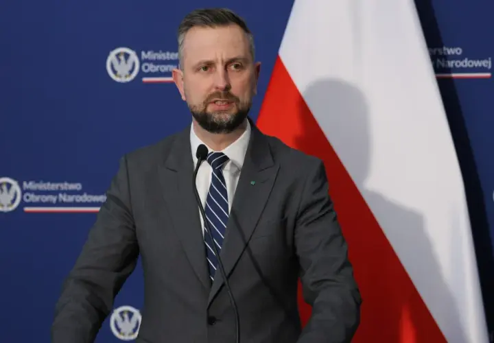 Mężczyzna w garniturze, minister obrony narodowej, przemawia przy mikrofonie na tle flagi Polski i logo ministerstwa.