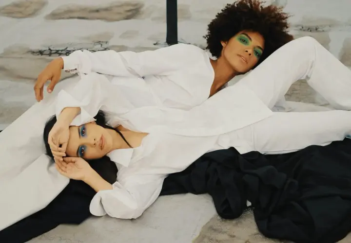 Two women in white shirts and pants, one with green eyeshadow, the other with blue. The fabric looks smooth, suggesting lyocell does not wrinkle easily.