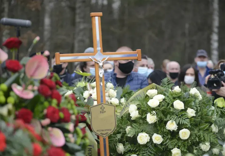 Drewniany krzyż z figurą Chrystusa na pogrzebie. Wieniec białych róż i czerwonych kwiatów. Ludzie w maseczkach. Co zrobić z krzyżem po pogrzebie?