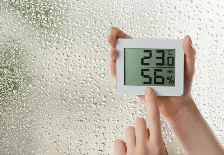 Manos sostienen un higrómetro digital que muestra 23.0°C y 56% de humedad, mientras gotas de lluvia cubren el cristal.