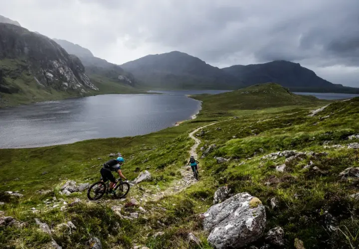 Two cyclists navigate a rocky trail with a vast lake and dramatic mountains behind them, showcasing the rugged beauty of Scotland bike tours.