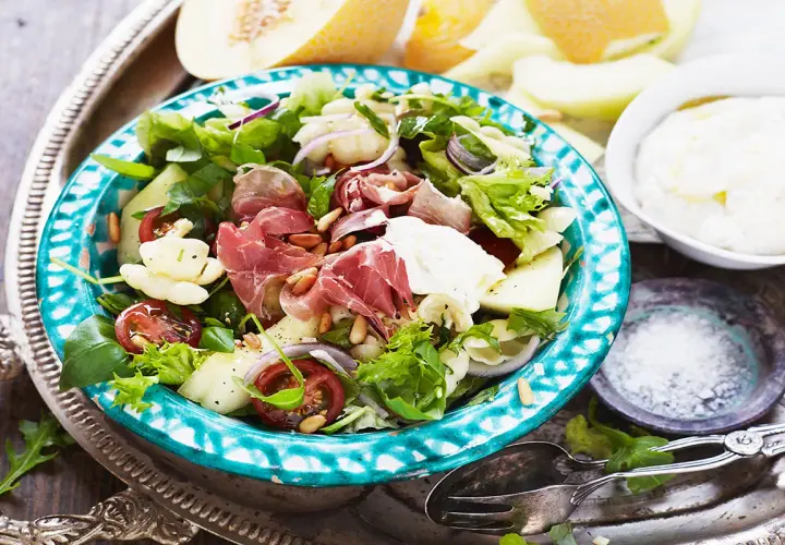 En härlig ost och skinksallad med melon, tomater och pinjenötter. Serveras på en vacker turkosblå skål.